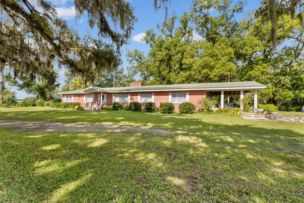A single-story red brick house with a metal roof and a carport sits on grass — 18951 SE 15TH STREET, WILLISTON, FL