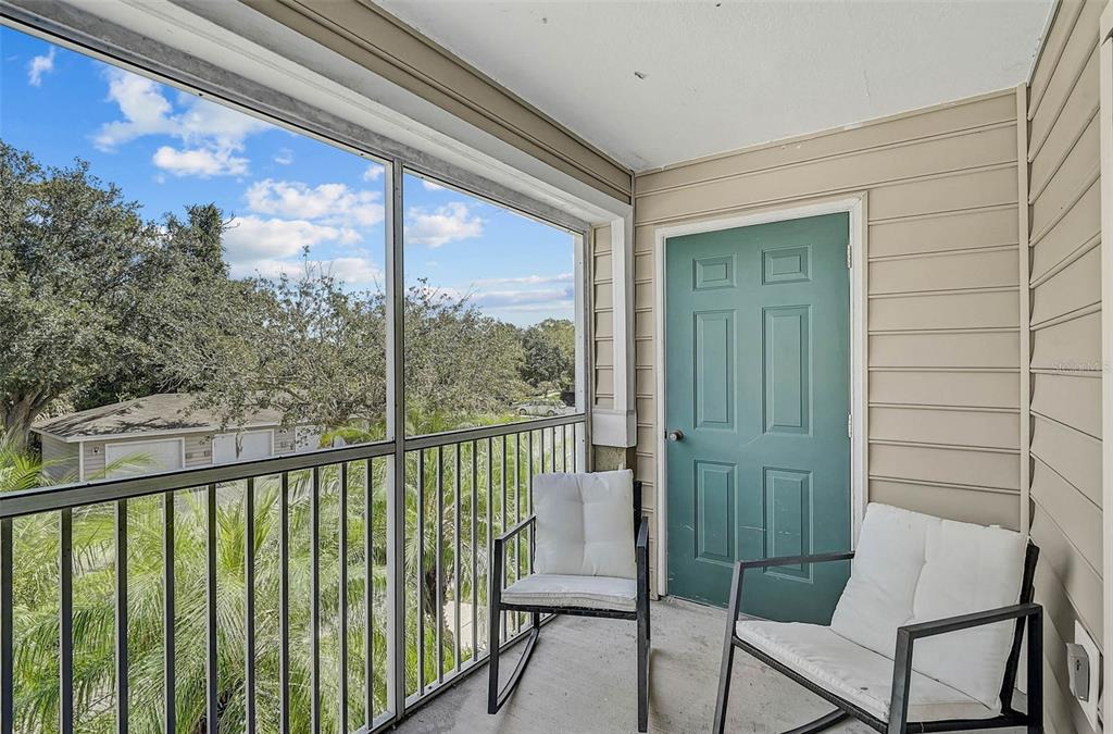 A screened-in balcony features two chairs, a teal door, and views of trees and buildings — 8827 MANOR LOOP UNIT 204, LAKEWOOD RANCH, FL