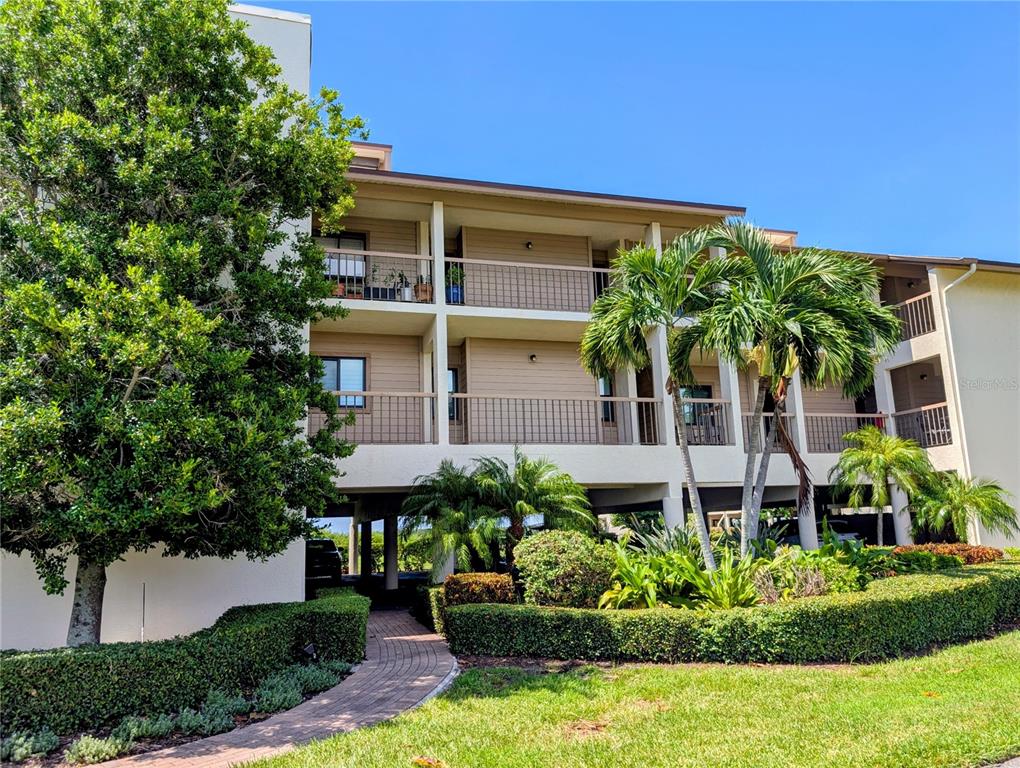 A multi-story apartment building with balconies and palm trees sits above a covered parking area — 3850 MARINERS WALK UNIT 723, CORTEZ, FL
