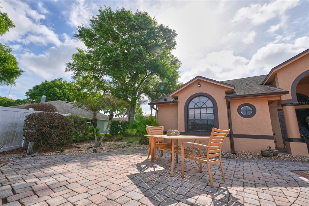 A paved patio with a dining set sits beside a tan house with a large tree — 103 OLYMPUS DRIVE, OCOEE, FL