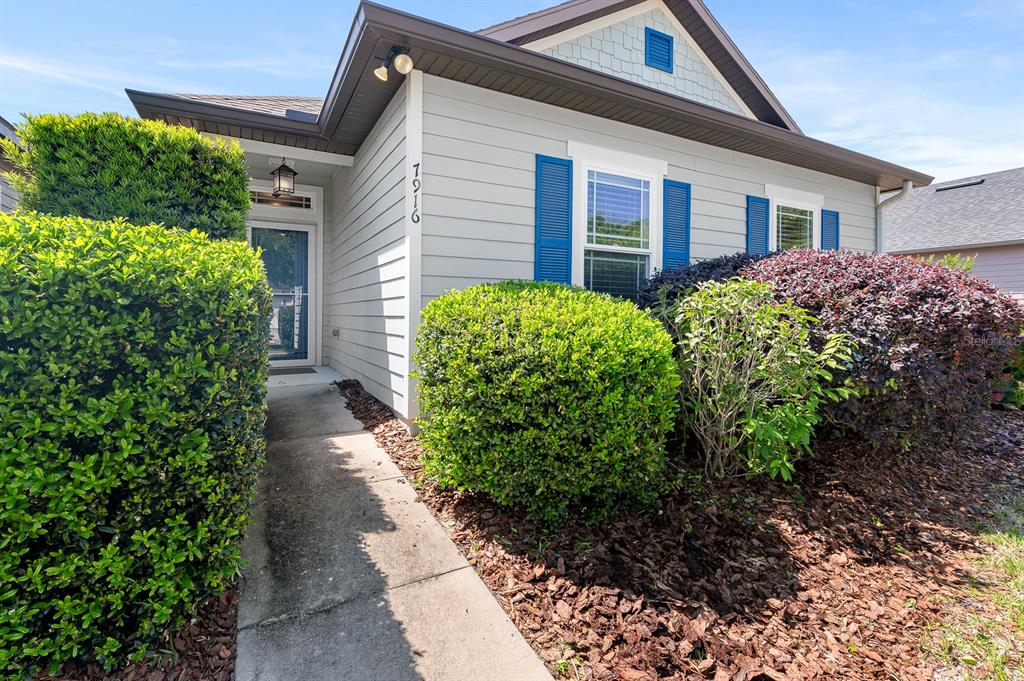 A light gray house exterior features blue shutters, a concrete walkway, and manicured landscaping — 7916 SW 80TH DRIVE, GAINESVILLE, FL