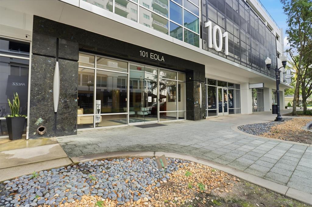 The exterior entrance of a modern building features glass doors, stone cladding, and landscaping — 101 S EOLA DRIVE UNIT 1104, ORLANDO, FL