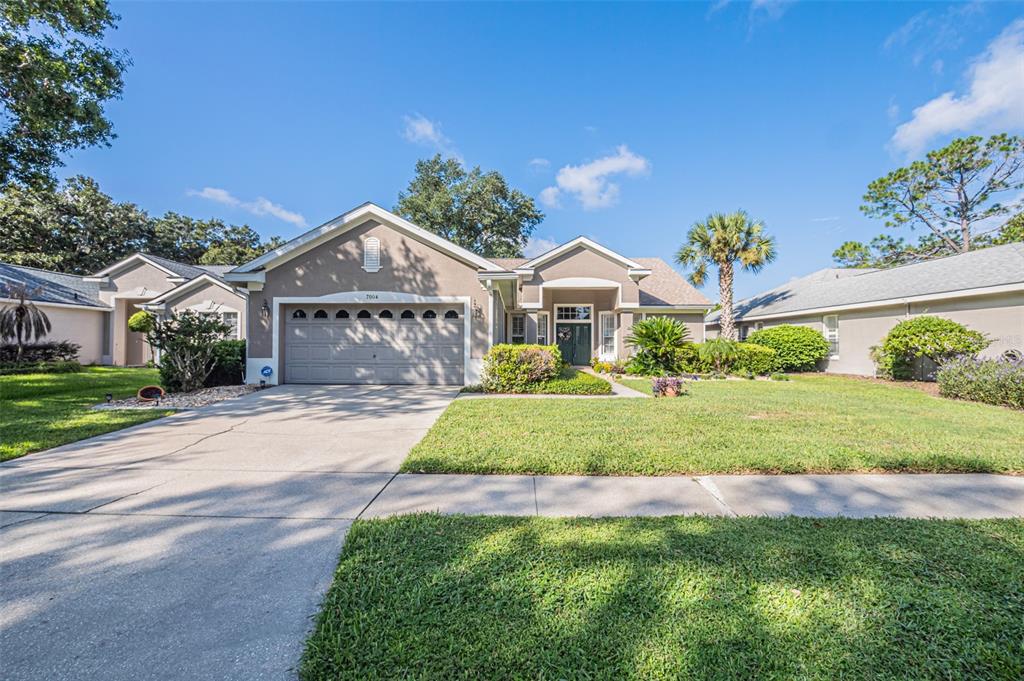 A single-story suburban house with a tan stucco exterior, a two-car garage, and a lawn — 7004 PINE HOLLOW DRIVE, MOUNT DORA, FL