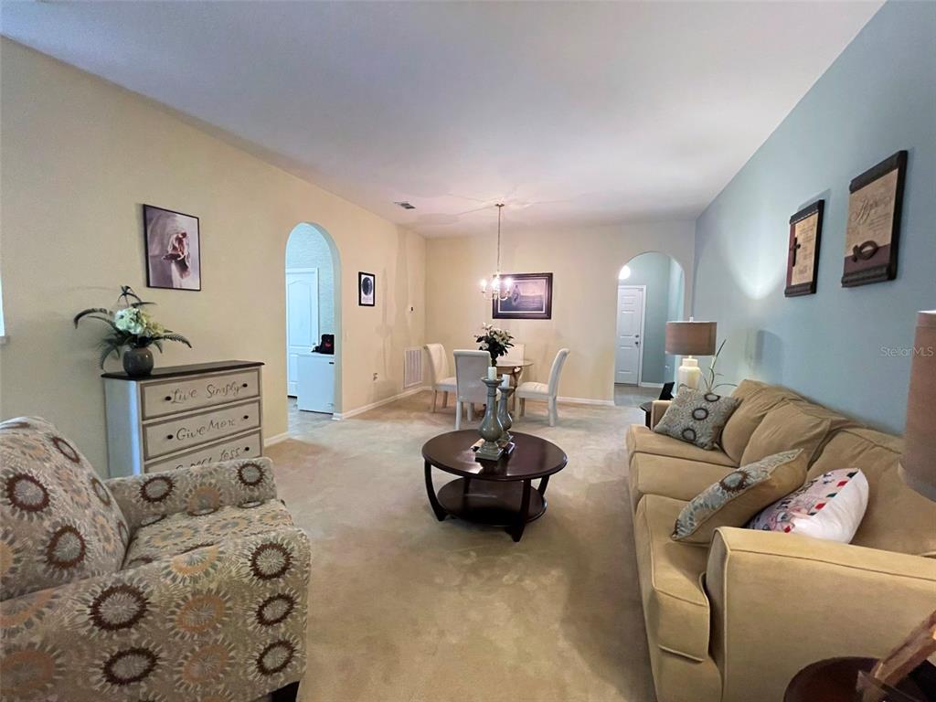 A living room with beige carpet, a tan sofa, patterned armchair, and a dining area beyond — 5804 DORNICH DRIVE, AUBURNDALE, FL