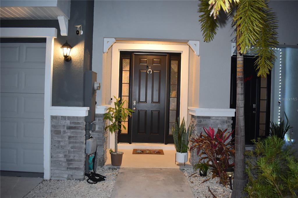 A dark front door with sidelights is flanked by potted plants and stone accents — 6572 SERENITY FALL LANE, SARASOTA, FL
