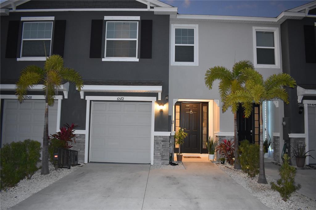 A two-story townhome features a gray garage, stone accents, and a front entrance with palms — 6572 SERENITY FALL LANE, SARASOTA, FL