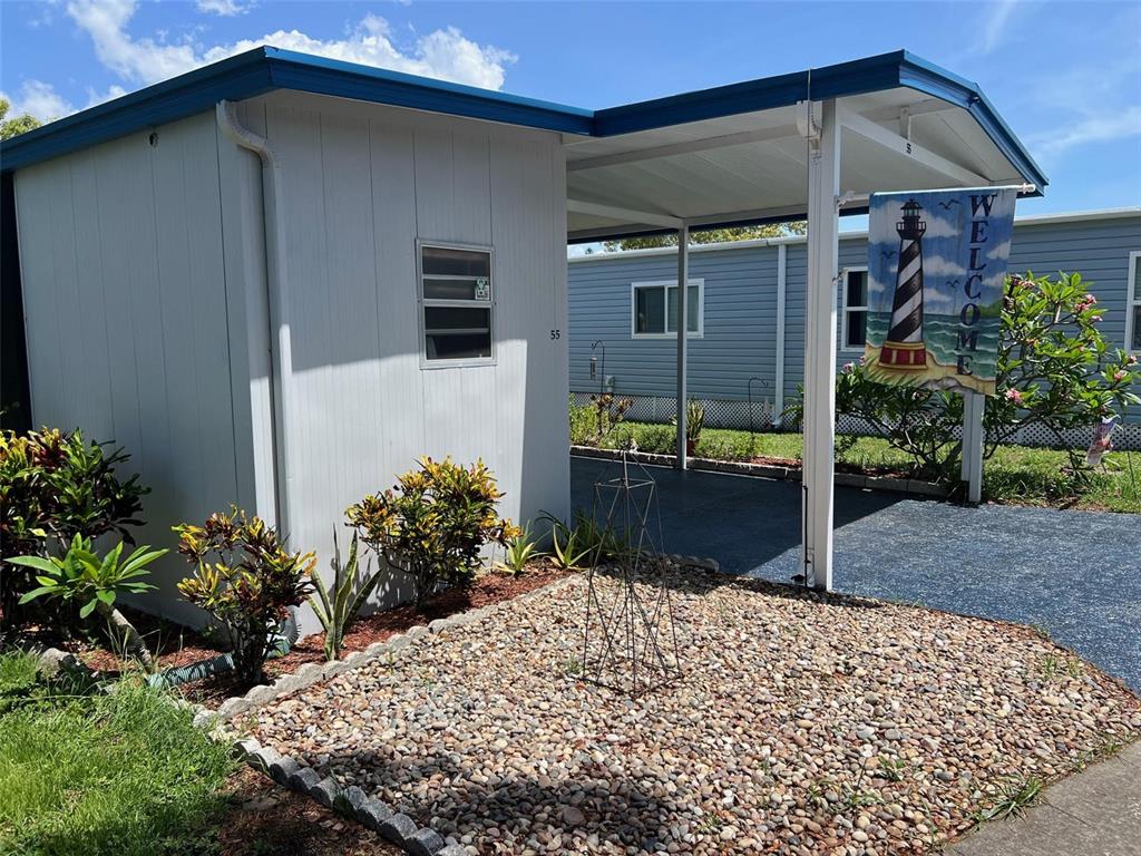A light-colored mobile home features a gravel front yard, a carport, and a welcome flag — 55 PALM FOREST DRIVE, LARGO, FL