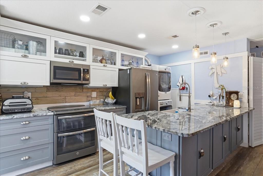 A kitchen features gray lower cabinets, white upper cabinets, a granite island, and wood-plank backsplash — 8931 BLIND PASS ROAD UNIT 360, ST PETE BEACH, FL