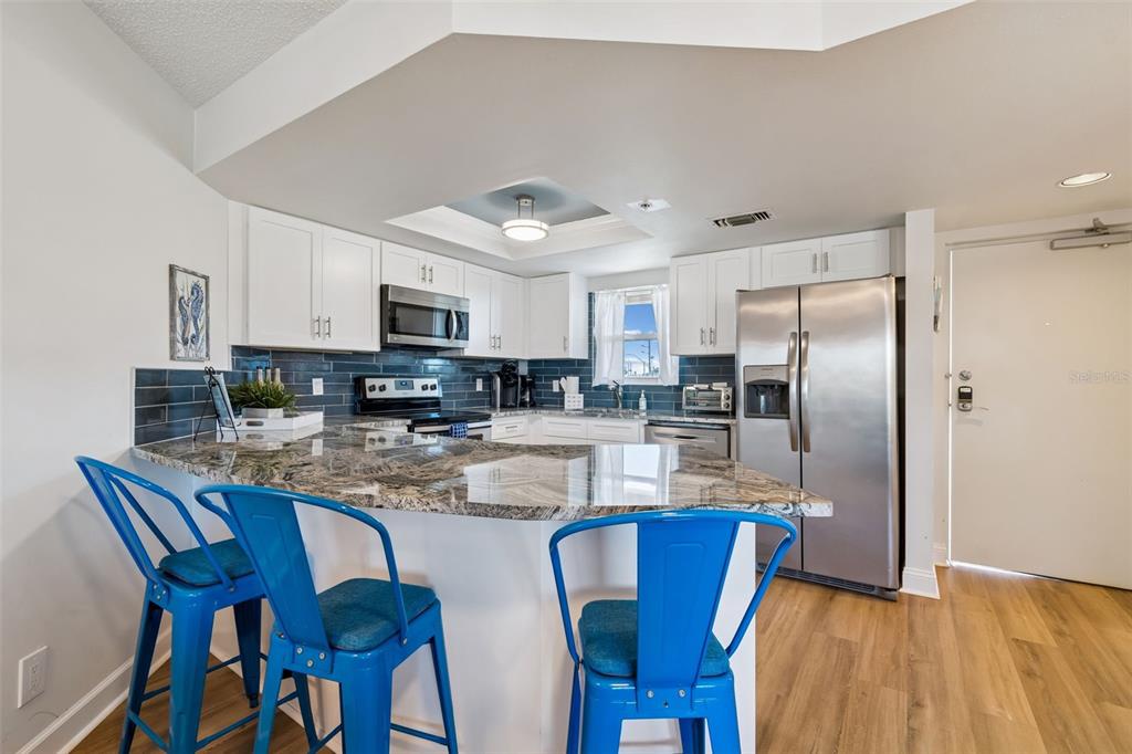 A kitchen features white cabinets, granite countertops, stainless steel appliances, and three blue metal stools — 4510 GULF BOULEVARD UNIT 308, ST PETE BEACH, FL