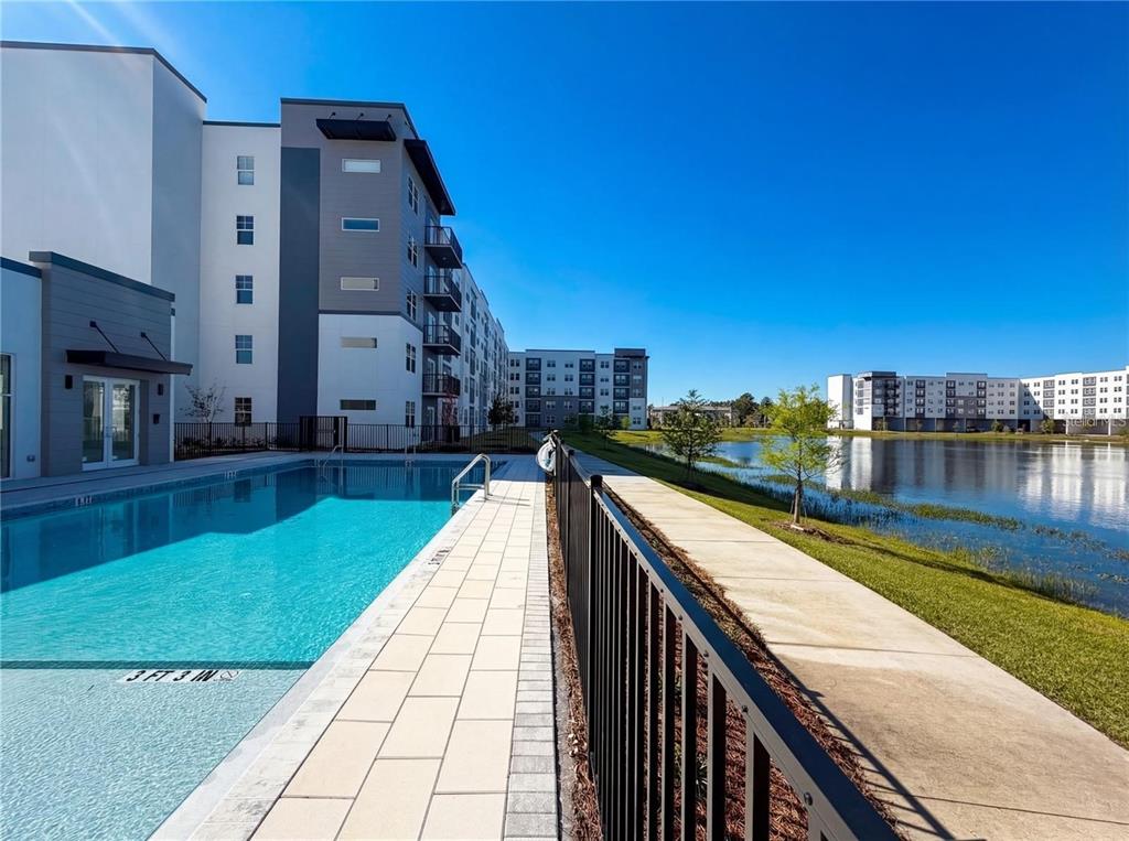 A swimming pool sits beside a modern apartment building and a lake under a clear sky — 5579 MILLENIA BOULEVARD UNIT 515, ORLANDO, FL