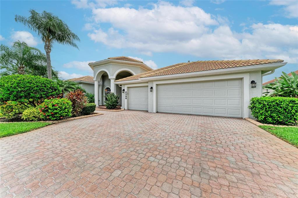 A single-story house with a paved driveway, three-car garage, and lush landscaping under a blue sky — 6988 74TH STREET CIRCLE E, BRADENTON, FL