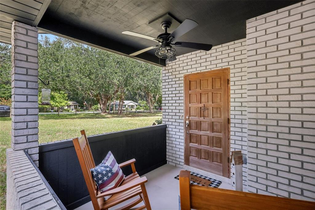 A covered front porch features a wooden rocking chair, a ceiling fan, and white brick walls — 209 JASPER STREET, BUSHNELL, FL