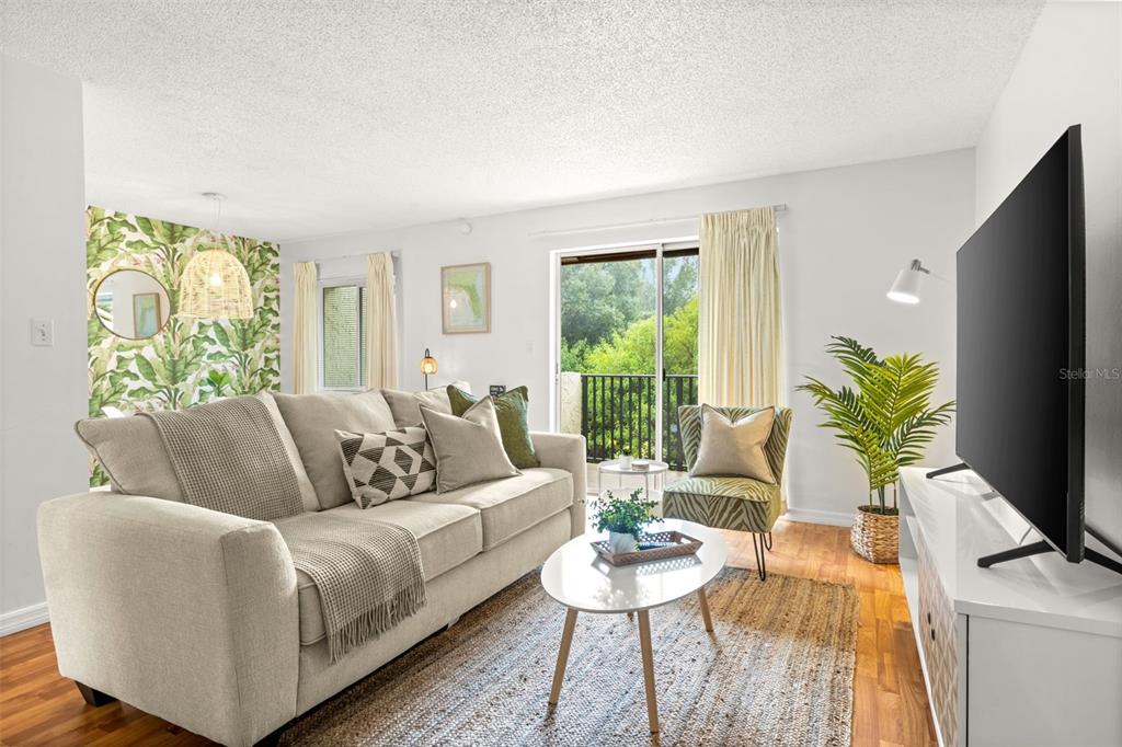 A living room features a beige sofa, a coffee table, a television, and tropical wallpaper — 4215 E BAY DRIVE UNIT 1806D, CLEARWATER, FL