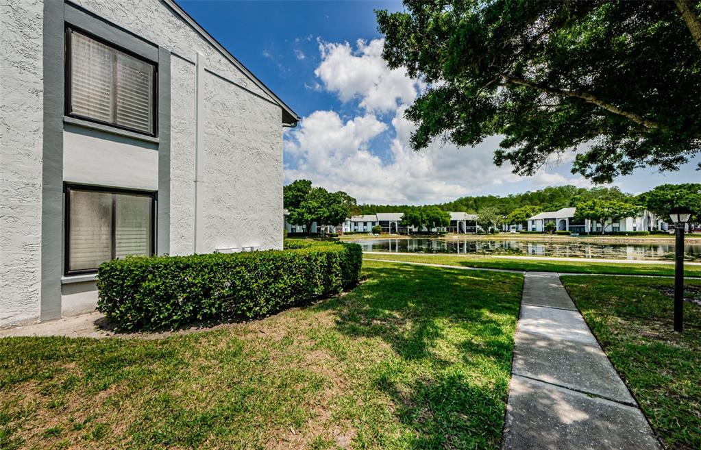 A white apartment building exterior sits next to a sidewalk, green lawn, and a pond — 1151 PINE RIDGE CIRCLE W UNIT F1, TARPON SPRINGS, FL