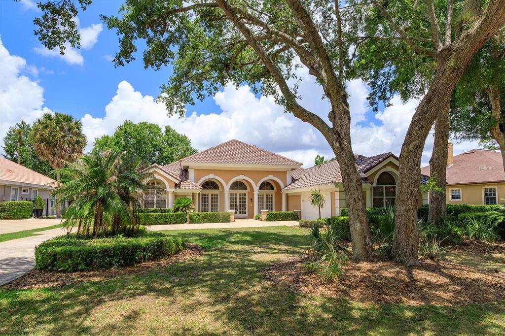 A tan single-story house with a tiled roof and arched windows sits behind large trees — 4965 SHORELINE CIRCLE, SANFORD, FL