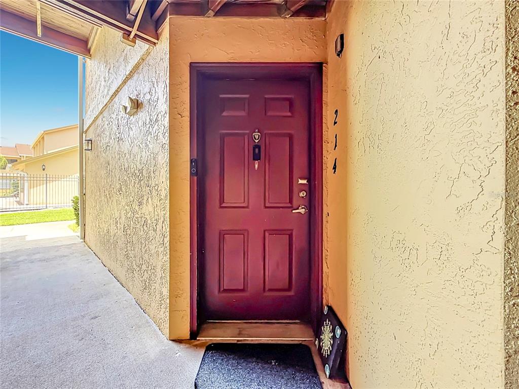 A dark red front door with a peephole and handle is set in a stucco wall — 1550 S BELCHER ROAD UNIT 214, CLEARWATER, FL