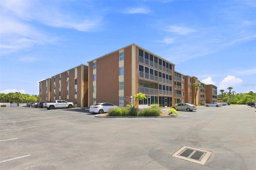 A multi-story apartment building with screened balconies stands behind a large paved parking lot — 12760 INDIAN ROCKS ROAD UNIT 1017, LARGO, FL