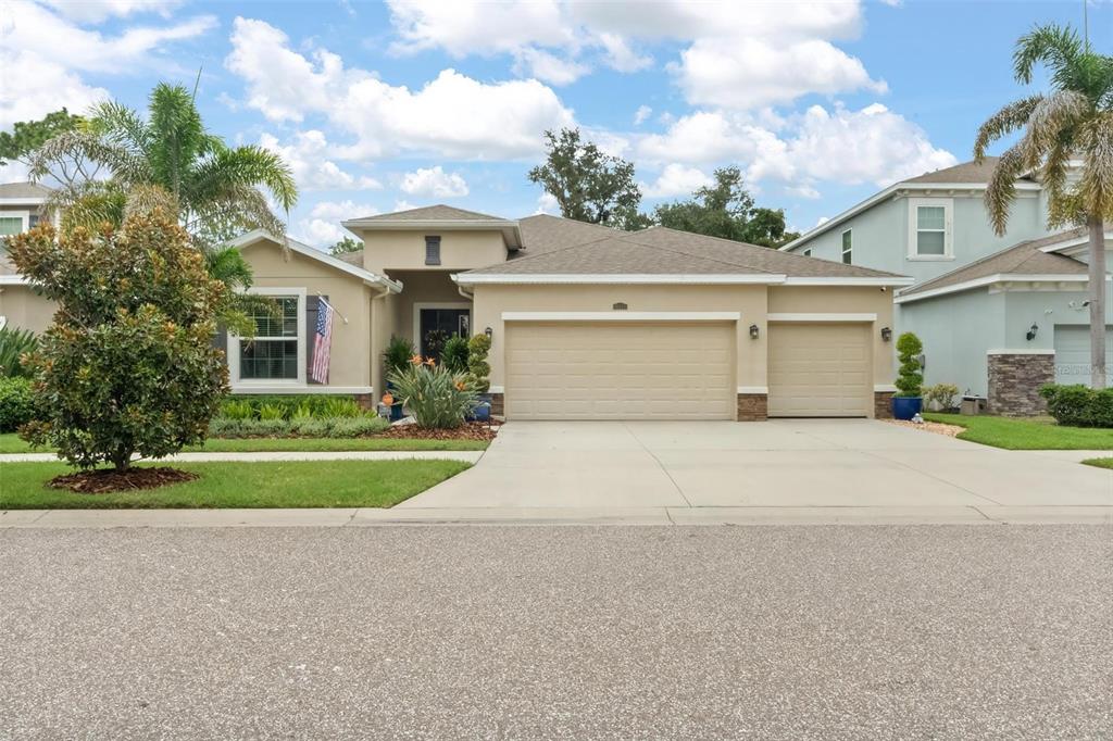 A single-story tan house with a three-car garage and an American flag near the entrance — 10321 RIVERDALE RISE DRIVE, RIVERVIEW, FL