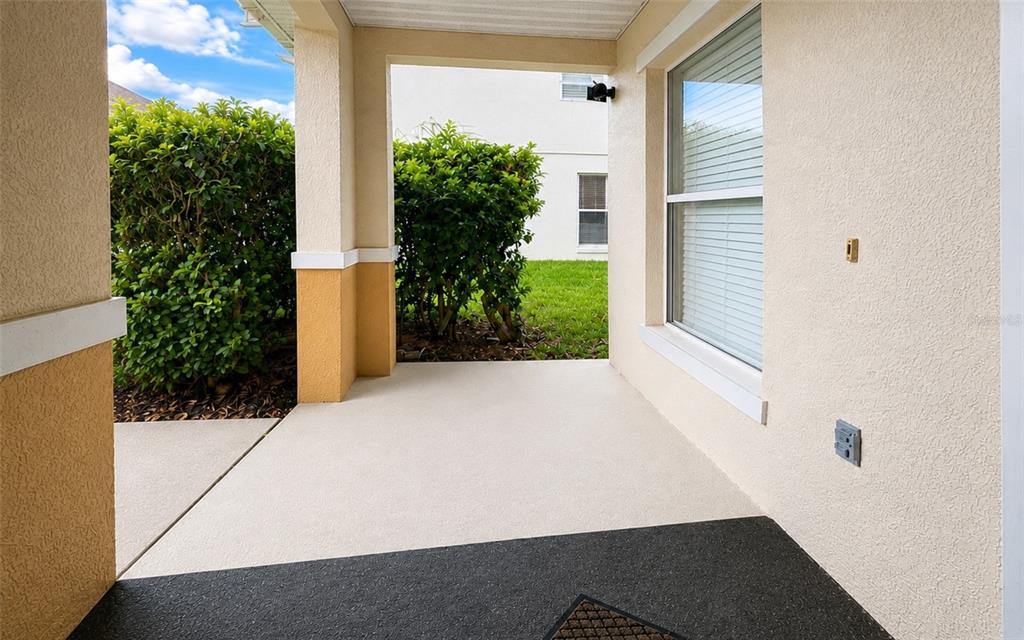 A covered concrete patio features a beige stucco wall, a window, and a nearby hedge — 133 RONA LANE, DAVENPORT, FL