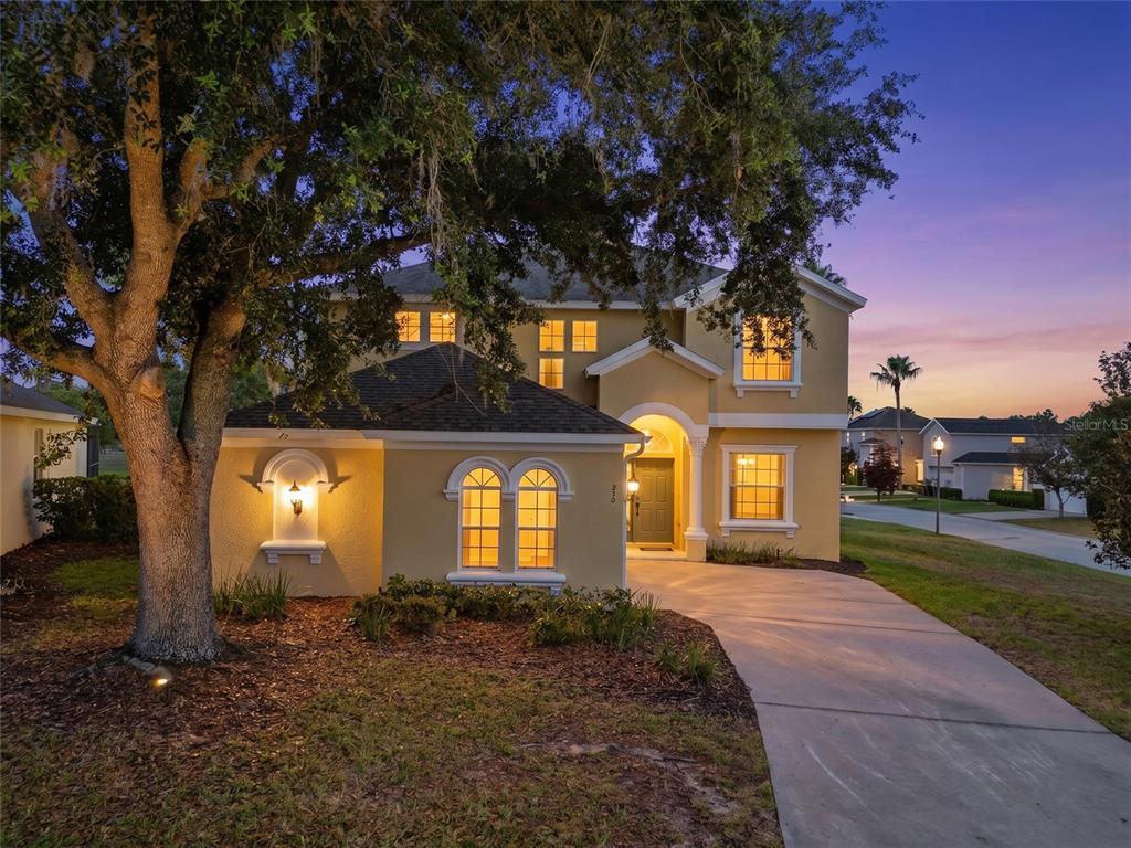 A two-story yellow stucco house with a driveway and a large tree at dusk — 353 PLUMOSO LOOP, DAVENPORT, FL