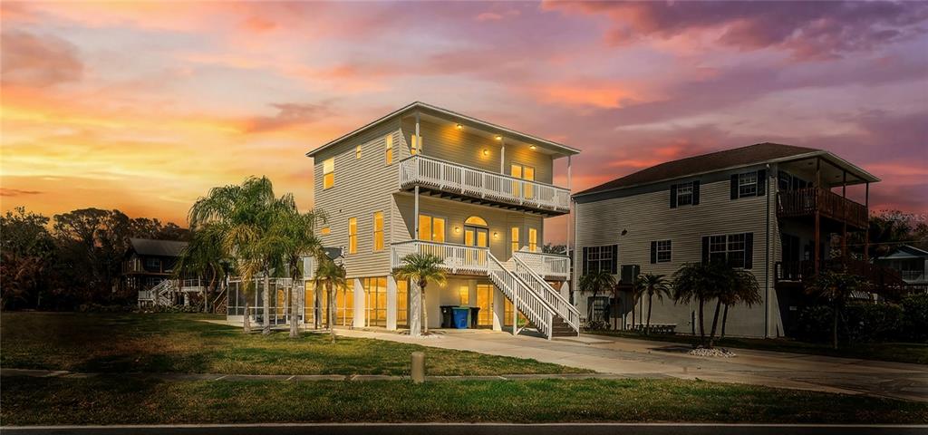 A three-story house with elevated decks and stairs stands against a sunset sky — 402 SHORE DRIVE W, OLDSMAR, FL