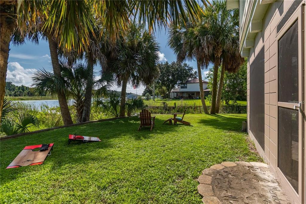 A grassy backyard features a cornhole game, two wooden chairs, and palm trees by water — 3 SE OCALE WAY, SUMMERFIELD, FL