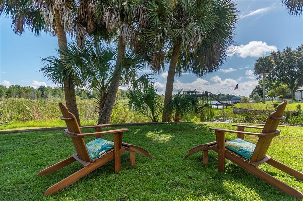 Two wooden lounge chairs sit on a grassy lawn overlooking a lake with palm trees — 3 SE OCALE WAY, SUMMERFIELD, FL