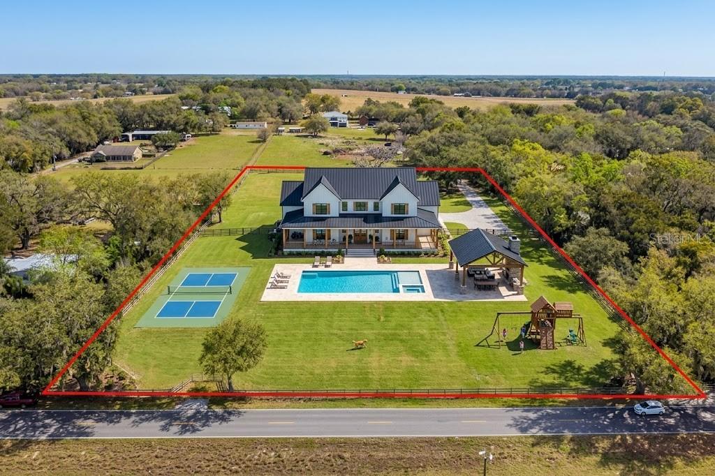 An aerial view shows a modern farmhouse with a pool, tennis court, and playground — 8302 WIRE ROAD, ZEPHYRHILLS, FL