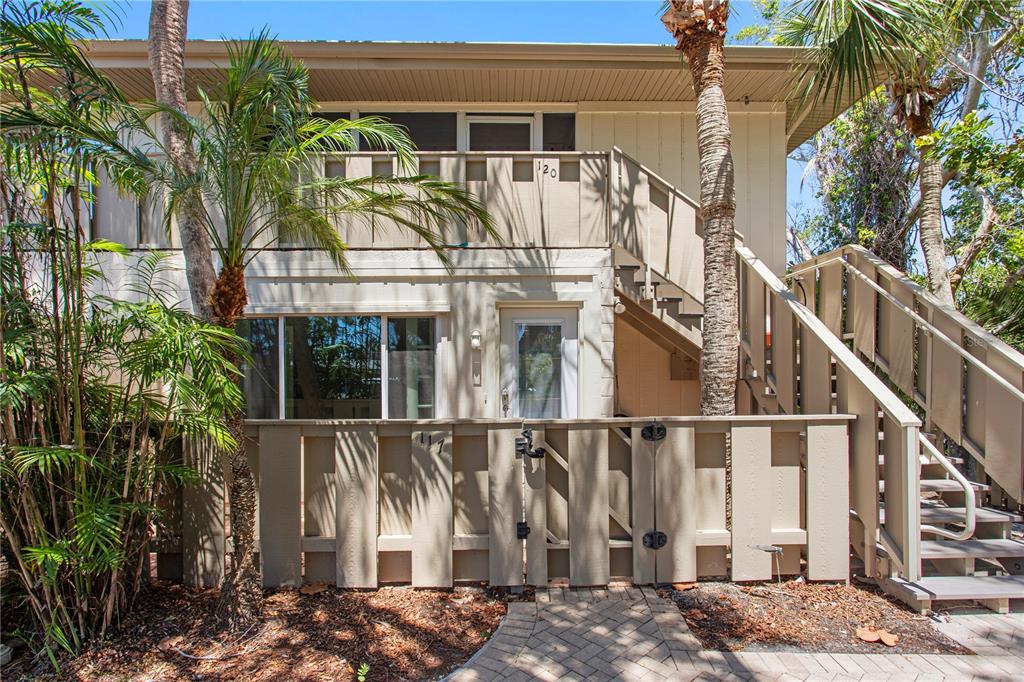A two-story exterior view of a tan building with a fenced patio and wooden stairs — 6700 GULF OF MEXICO DRIVE UNIT 117, LONGBOAT KEY, FL