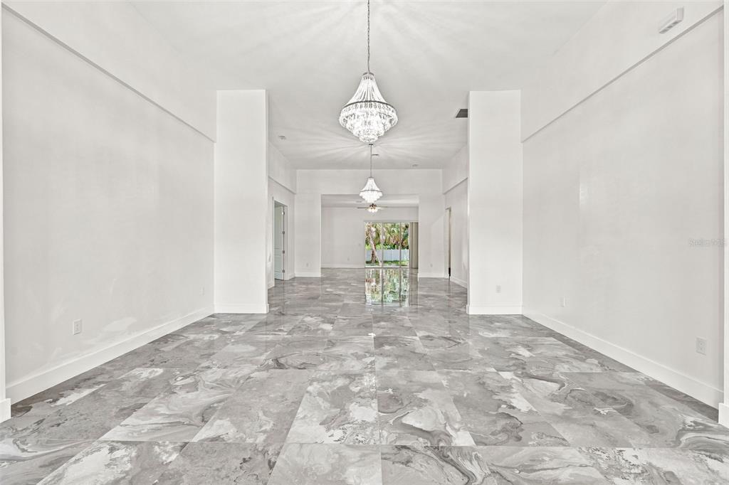 A spacious, empty room featuring white walls, two crystal chandeliers, and gray marble-patterned tile flooring — 1853 DELLA DRIVE, NAPLES, FL