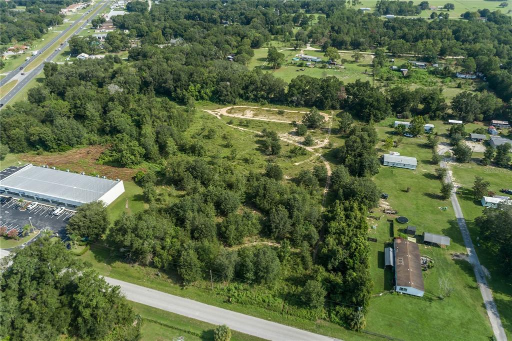 An aerial view shows a large, wooded vacant lot situated between a commercial building and residential properties — 7711 SE 126TH PLACE, BELLEVIEW, FL