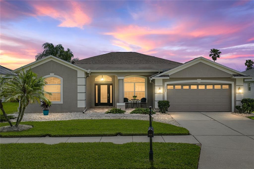 A single-story suburban house with a gray stucco exterior, a two-car garage, and a sunset sky — 1542 SWAMP ROSE LANE, TRINITY, FL