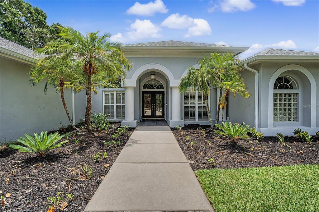 A concrete walkway leads to the arched entryway of a gray house with palm trees — 3692 JOHNATHON AVENUE, PALM HARBOR, FL