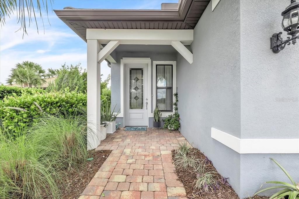 A paved walkway leads to the front entrance of a house with gray stucco walls — 7538 SUMMERLAND COVE, LAKEWOOD RANCH, FL