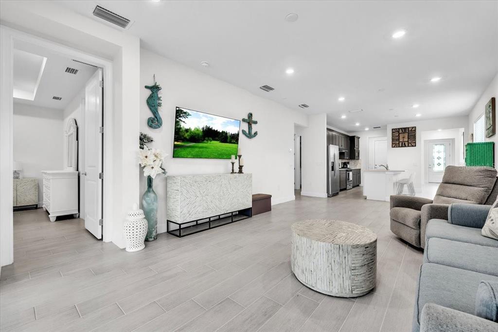 A bright living room features light wood-look tile flooring, a wall-mounted television, and an open kitchen — 7538 SUMMERLAND COVE, LAKEWOOD RANCH, FL