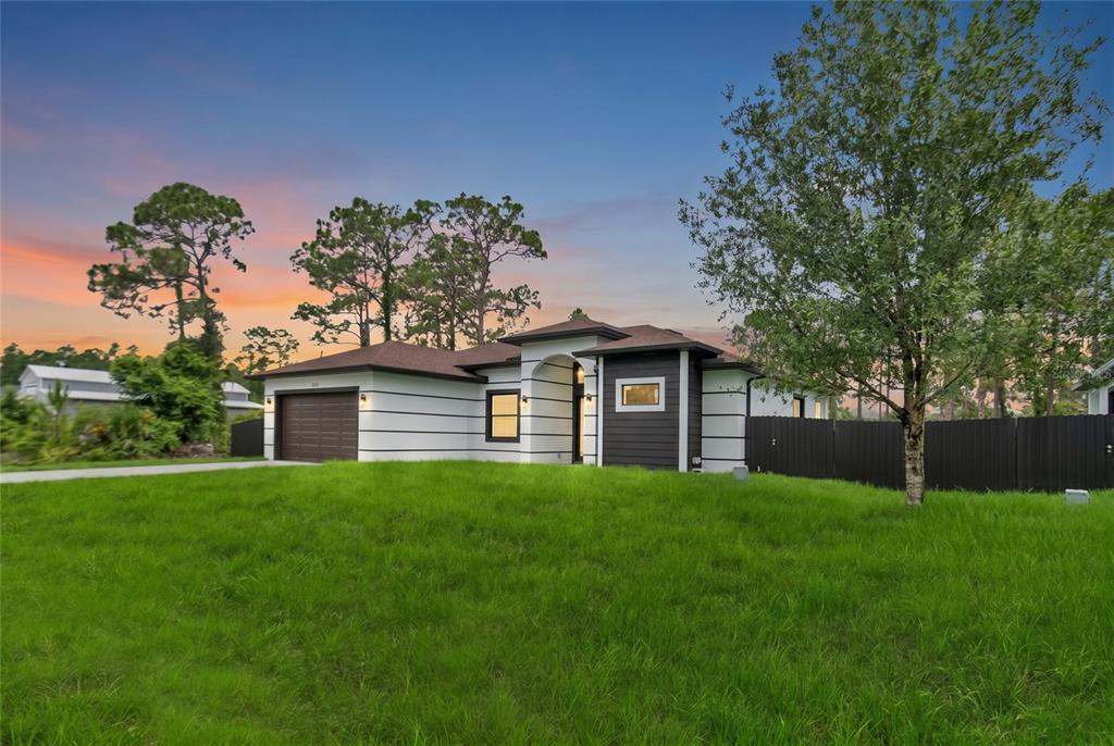 A single-story house with a two-car garage, white siding, and dark trim under a sunset sky — 1047 MERRY STREET E, LEHIGH ACRES, FL