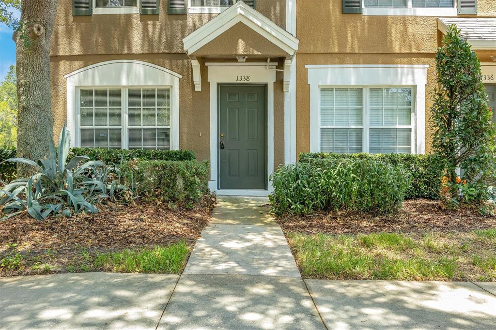A tan townhouse exterior features a dark green front door, white trim, and landscaped bushes — 1338 STANDRIDGE DRIVE, ZEPHYRHILLS, FL