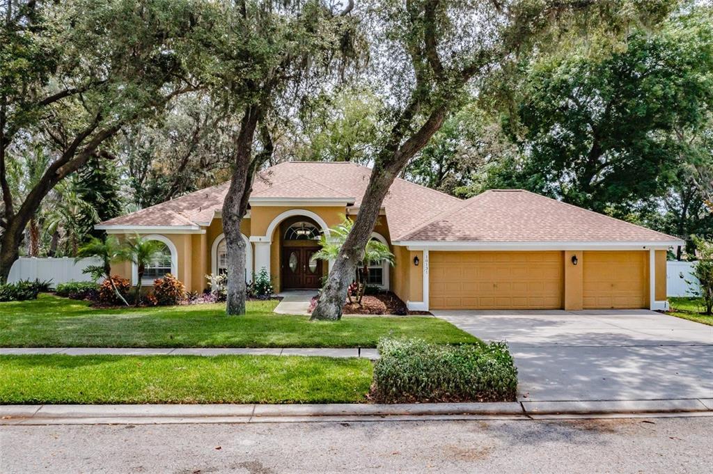 A single-story yellow house with a three-car garage and arched entryway sits behind mature trees — 10131 PADDOCK OAKS DRIVE, RIVERVIEW, FL