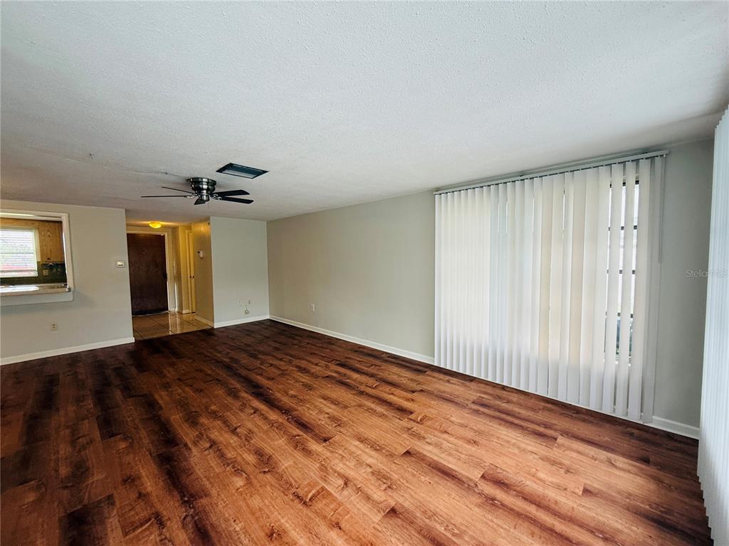 An empty living room features wood-look flooring, a ceiling fan, and large vertical window blinds — 7910 LILAC DRIVE, PORT RICHEY, FL