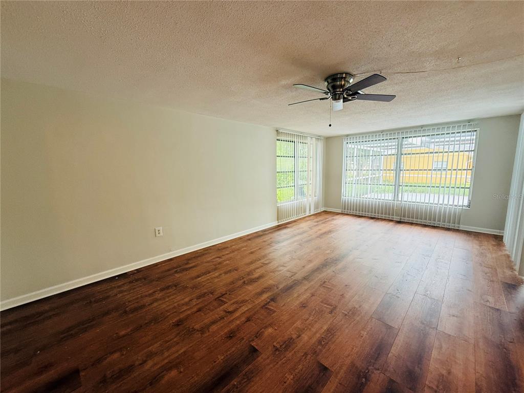 An empty living room features wood-look flooring, a ceiling fan, and large windows with blinds — 7910 LILAC DRIVE, PORT RICHEY, FL