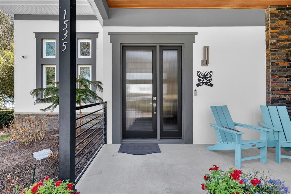 A modern front porch features a dark-framed glass door, two light blue chairs, and landscaping — 1535 SUNSET CIRCLE, MOUNT DORA, FL