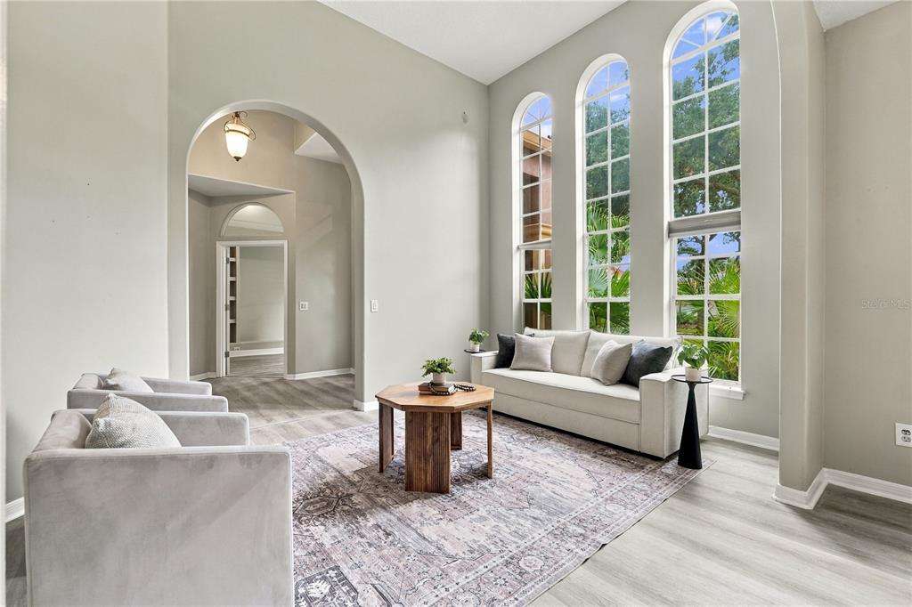 A bright living room features tall arched windows, light wood-look flooring, and a neutral sofa — 7628 MILANO DRIVE, ORLANDO, FL
