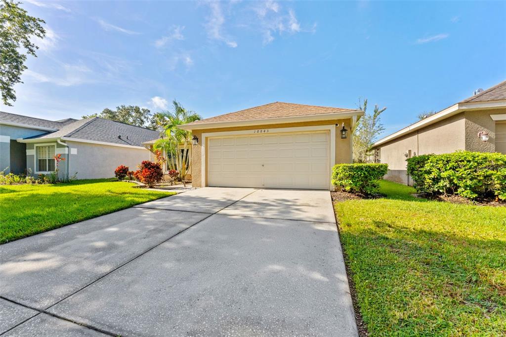 A single-story house with a beige stucco exterior, a two-car garage, and a concrete driveway — 18843 MAISONS DRIVE, LUTZ, FL