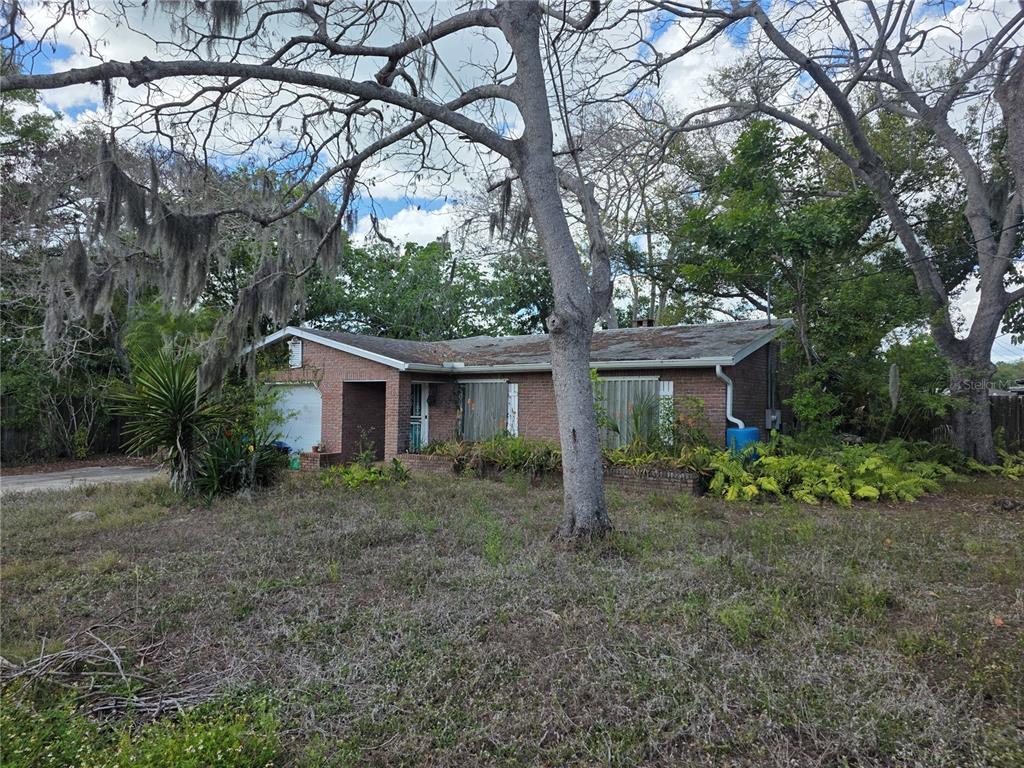 A single-story brick house with a garage and overgrown landscaping sits under large trees — 1016 GREER DRIVE, SARASOTA, FL