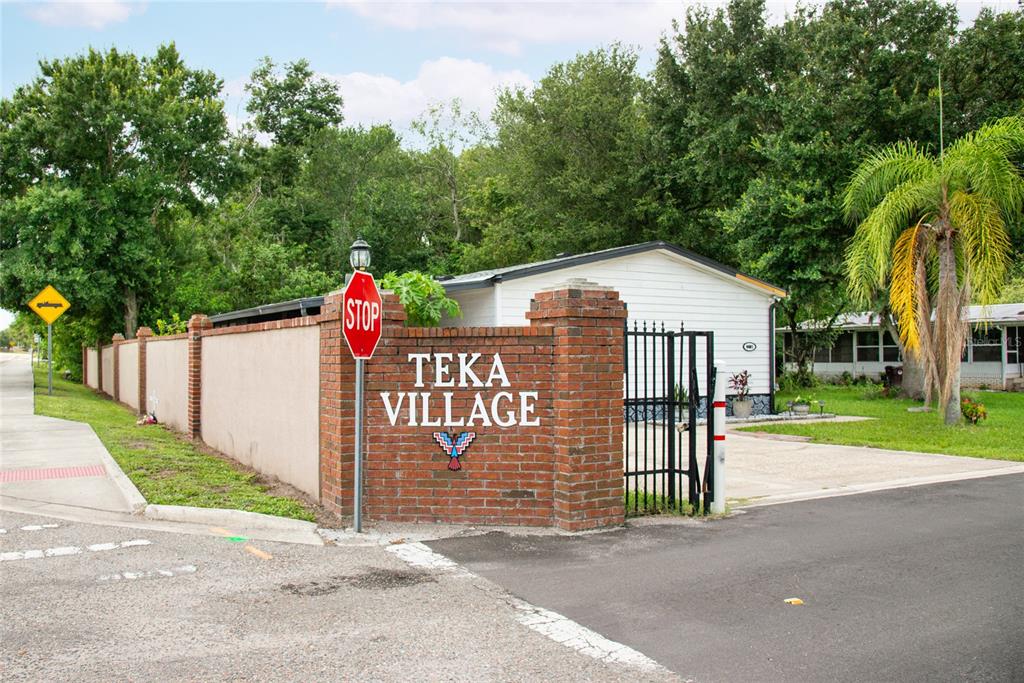 A brick entrance sign for Teka Village stands beside a gated driveway and white building — 2542 LONGPINE LANE, ST CLOUD, FL