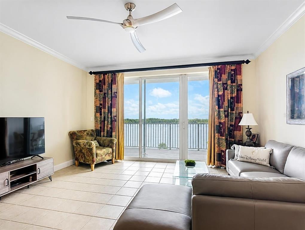 A living room features tile flooring, a grey sectional sofa, and a balcony overlooking water — 13427 BLUE HERON BEACH DRIVE UNIT 402, ORLANDO, FL