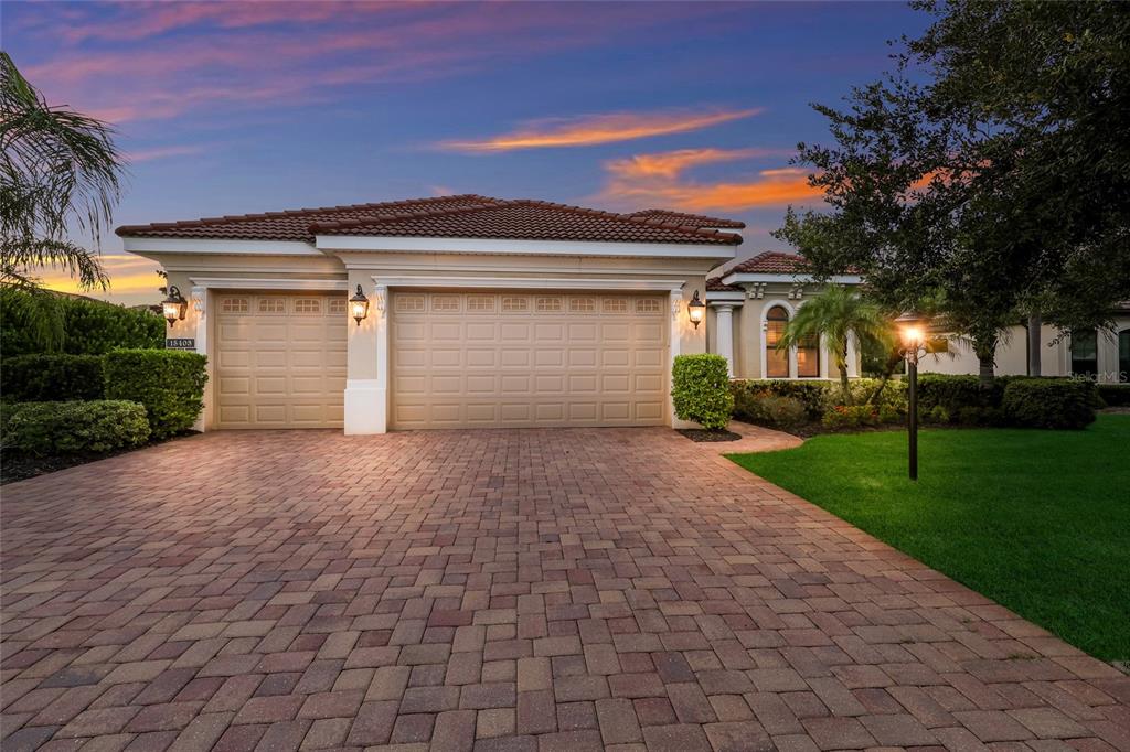 A beige house with a three-car garage and a paver driveway at sunset — 15403 HELMSDALE PLACE, LAKEWOOD RANCH, FL