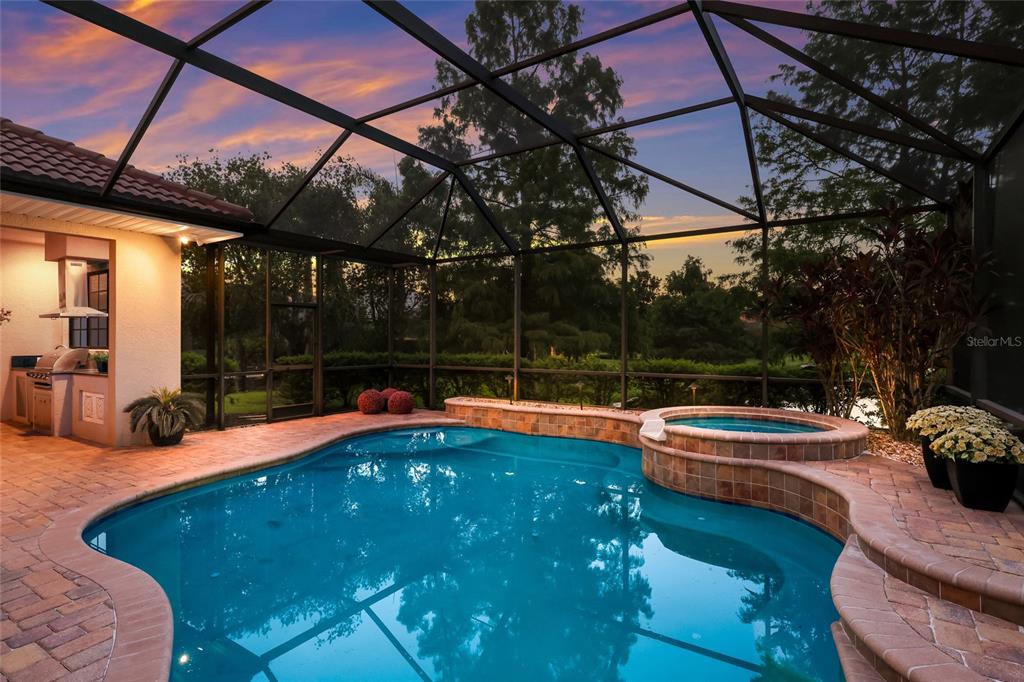 A screened-in pool and spa area with a brick patio and outdoor kitchen at sunset — 15403 HELMSDALE PLACE, LAKEWOOD RANCH, FL