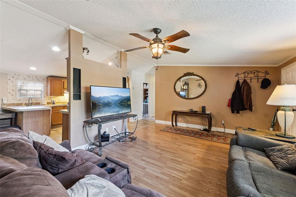 A living room with wood-look flooring, a ceiling fan, a television, and an adjacent kitchen — 680 WILD ACRES ROAD, OSTEEN, FL