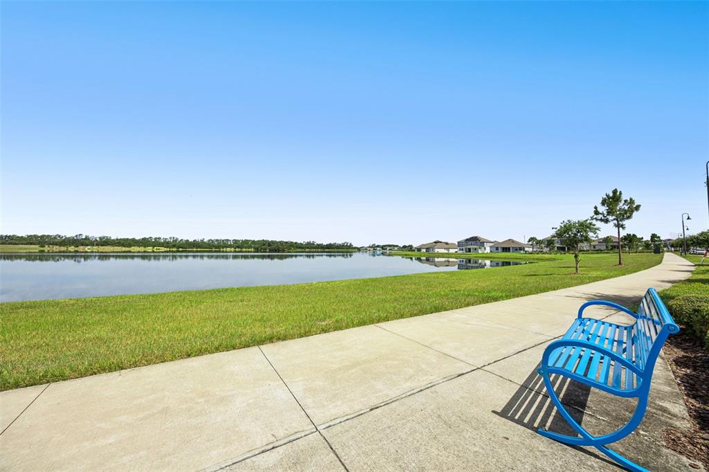 A blue metal bench sits on a concrete sidewalk beside a grassy lawn and lake — 2975 ADDISON BOULEVARD, ST CLOUD, FL